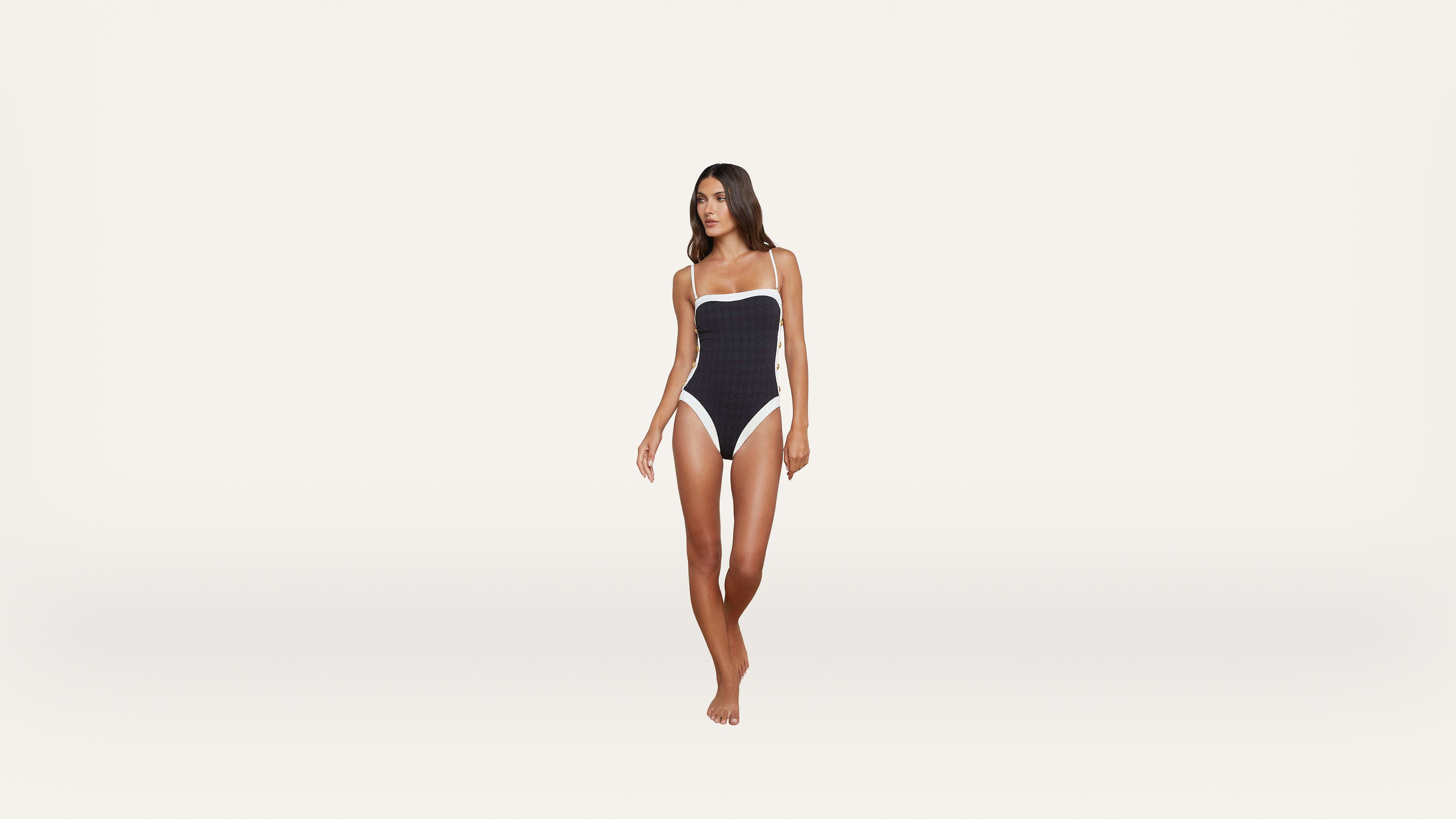A woman stands barefoot against a plain white background, wearing a navy blue one-piece swimsuit with white trim.