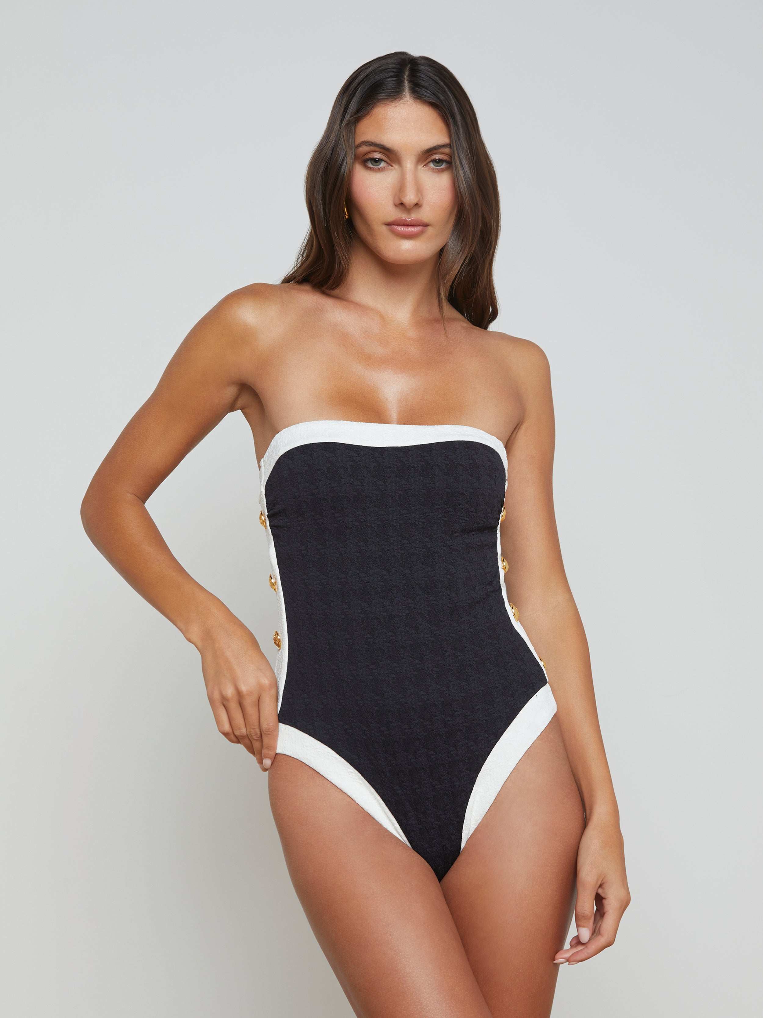 A woman models the Tory Bandeau One-Piece Swimsuit, featuring a strapless design with high-leg cut in black, accented by white trim and gold buttons, set against a plain light background.