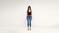 Woman standing against a plain white background, wearing sunglasses, a black tank top, blue jeans, a brown belt, and tan heels.