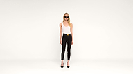 A woman wearing sunglasses, a white tank top, black pants with a belt, and black heels stands against a plain white background.