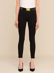 Indossa i jeans skinny a vita alta Margot Cropped in nero — un modello skinny realizzato in denim elasticizzato — abbinati a un top bianco senza maniche, una cintura nera con una grande fibbia dorata e tacchi neri a punta; la modella è in posa su uno sfondo beige a tinta unita, con solo la parte inferiore del corpo visibile.