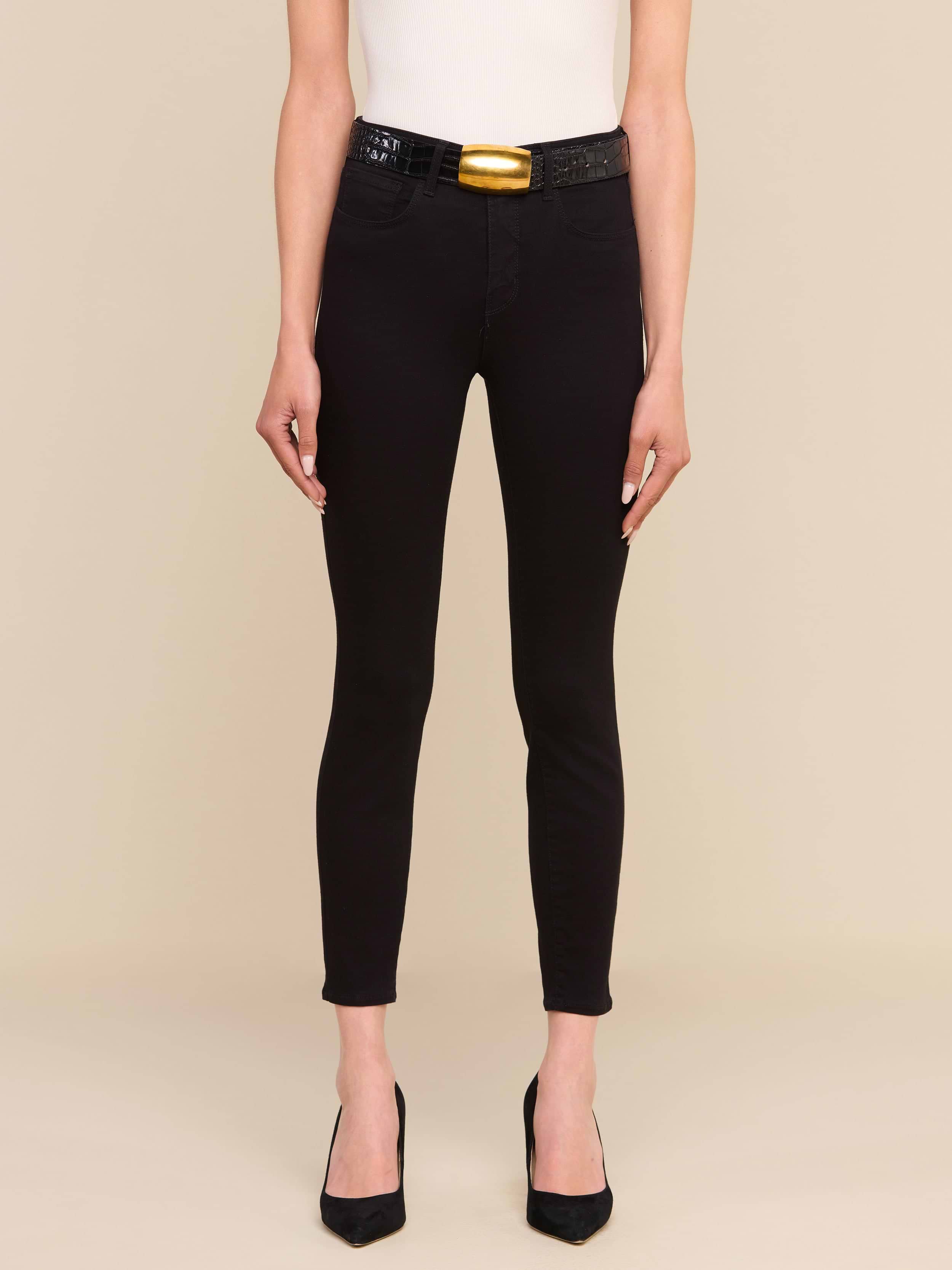 Indossa i jeans skinny a vita alta Margot Cropped in nero — un modello skinny realizzato in denim elasticizzato — abbinati a un top bianco senza maniche, una cintura nera con una grande fibbia dorata e tacchi neri a punta; la modella è in posa su uno sfondo beige a tinta unita, con solo la parte inferiore del corpo visibile.