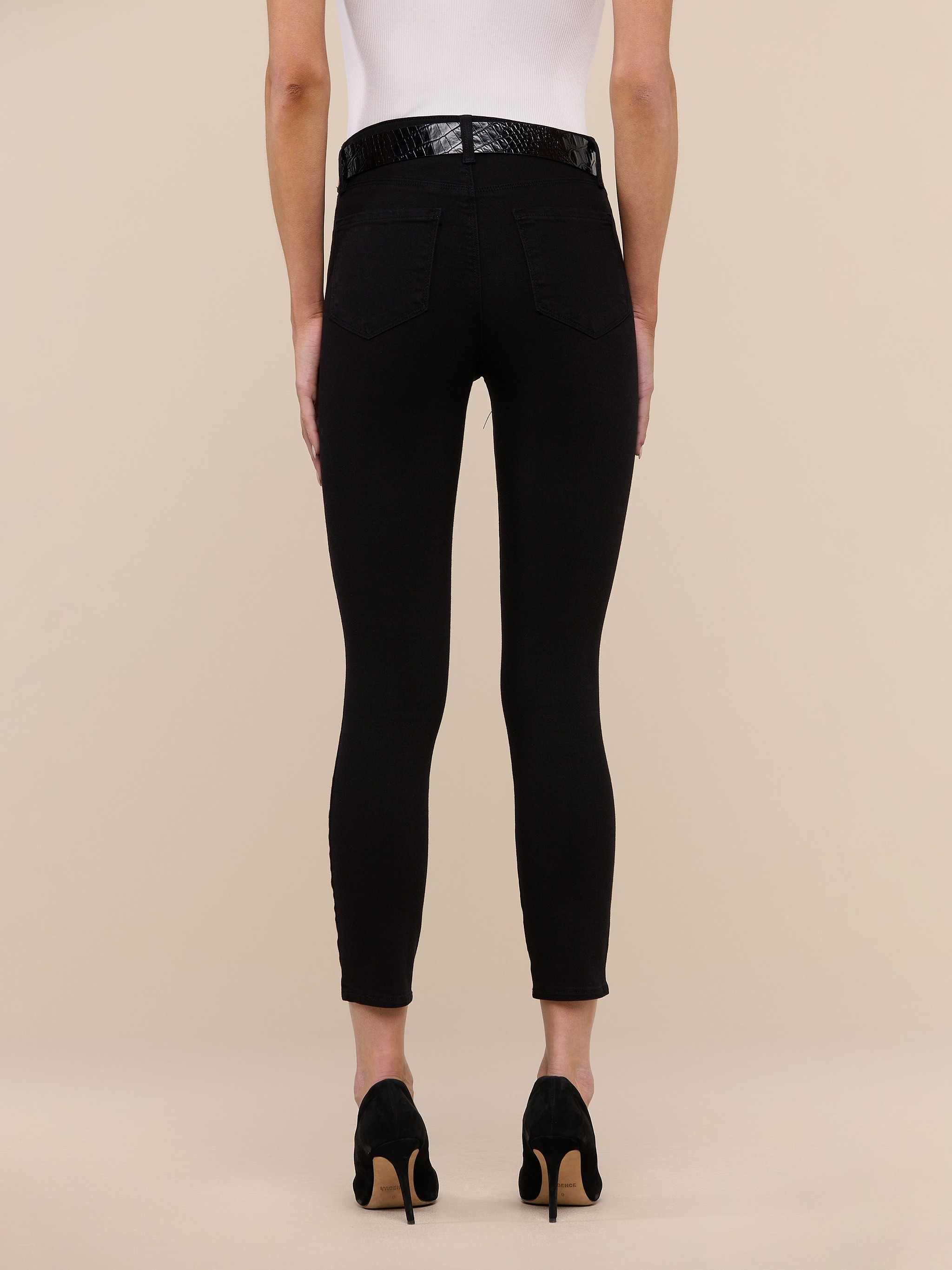 Person standing facing away, wearing a white top, the Margot Cropped Skinny Jean in black high-rise stretch denim, a black belt, and black high-heeled shoes against a plain white background.