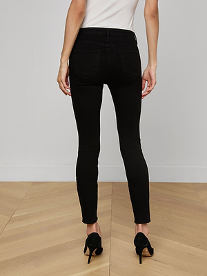 A person stands on a light wood floor, seen from the back, wearing Margot Cropped Skinny Jeans in black stretch denim, black high heels, and a white top.