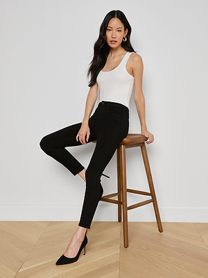 A woman with long dark hair, in a white sleeveless top and black Margot Cropped Skinny Jeans with black heels, sits sideways on a wooden stool against a plain white wall and light wooden floor.