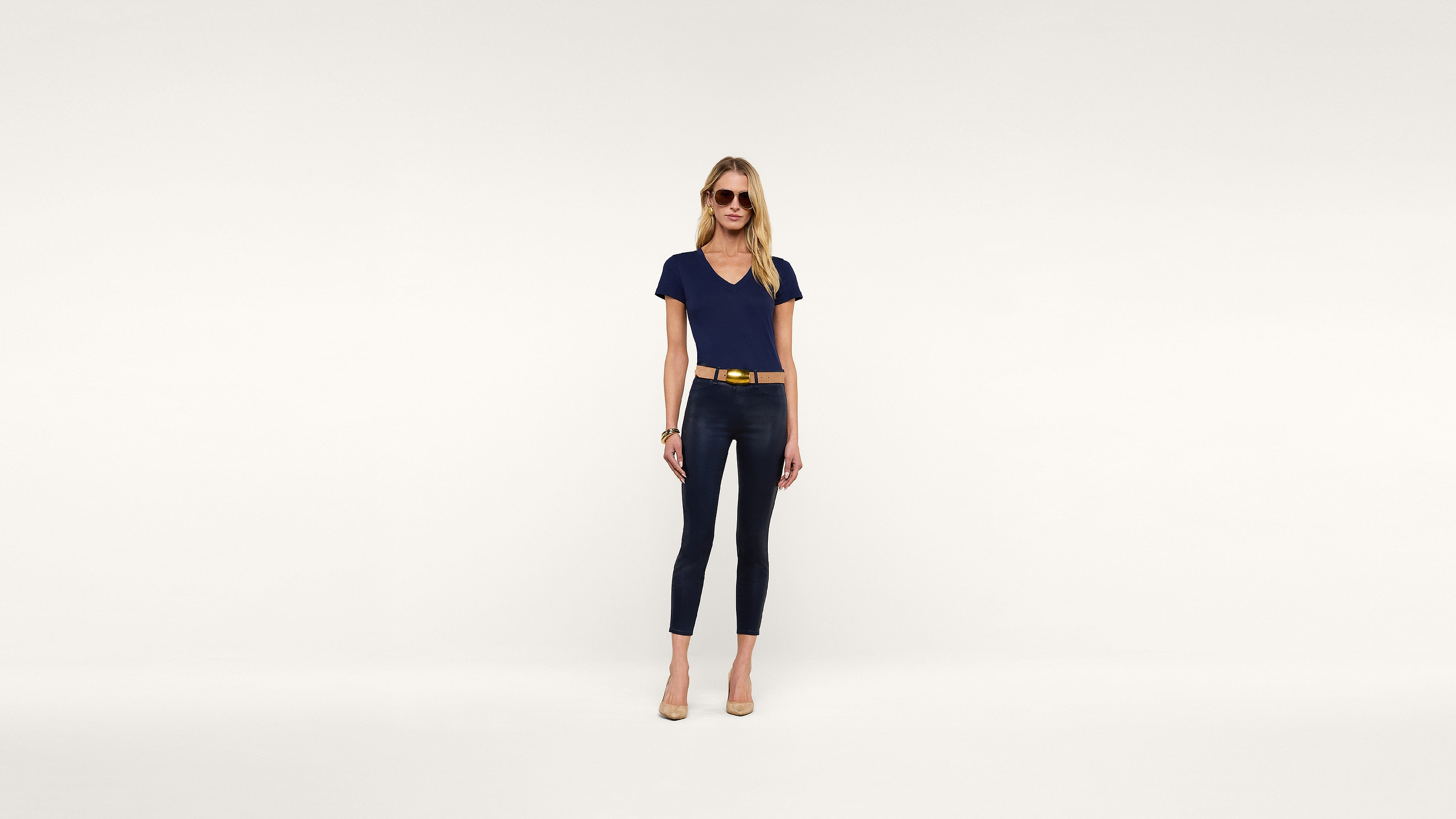 A woman wearing sunglasses, a navy blue t-shirt, dark blue cropped pants, a yellow belt, and beige heels poses confidently against a plain white background.