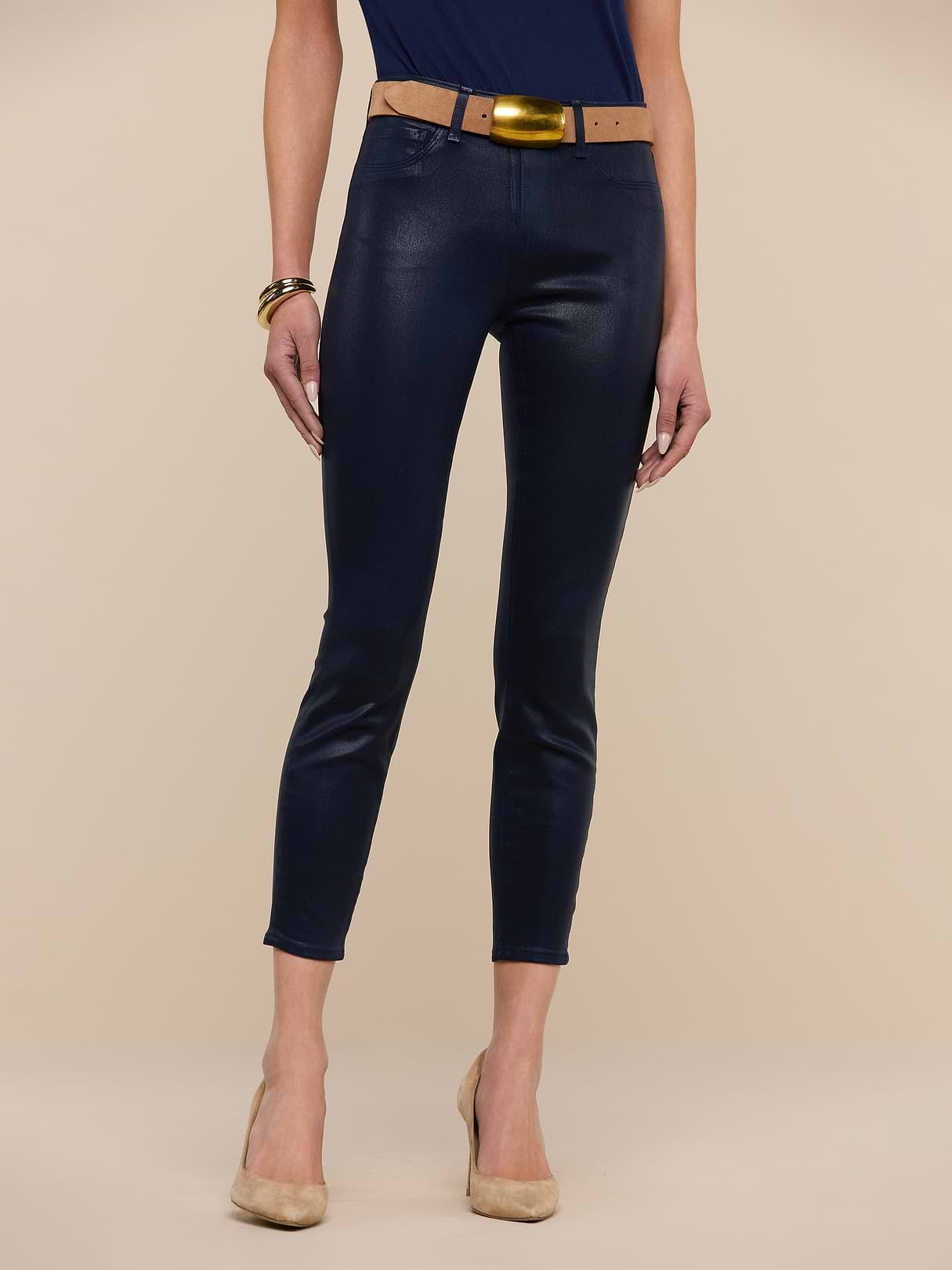 A person wears Margot Coated Cropped Skinny Jeans, a contoured waistband, a tan belt with gold buckle, navy top, tan heels, and gold bangles while standing against a plain beige background.