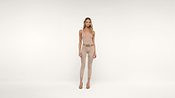 A woman in a beige sleeveless top, matching pants, and heels stands against a plain white background, looking straight ahead with a neutral expression.