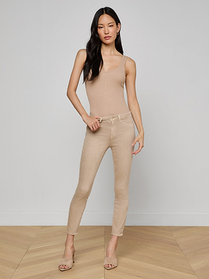 A woman stands indoors against a light gray wall, wearing a beige sleeveless bodysuit and Margot Cropped Skinny Jeans with a contoured waistband. She has long dark hair, heeled sandals, and rests one hand on her hip.