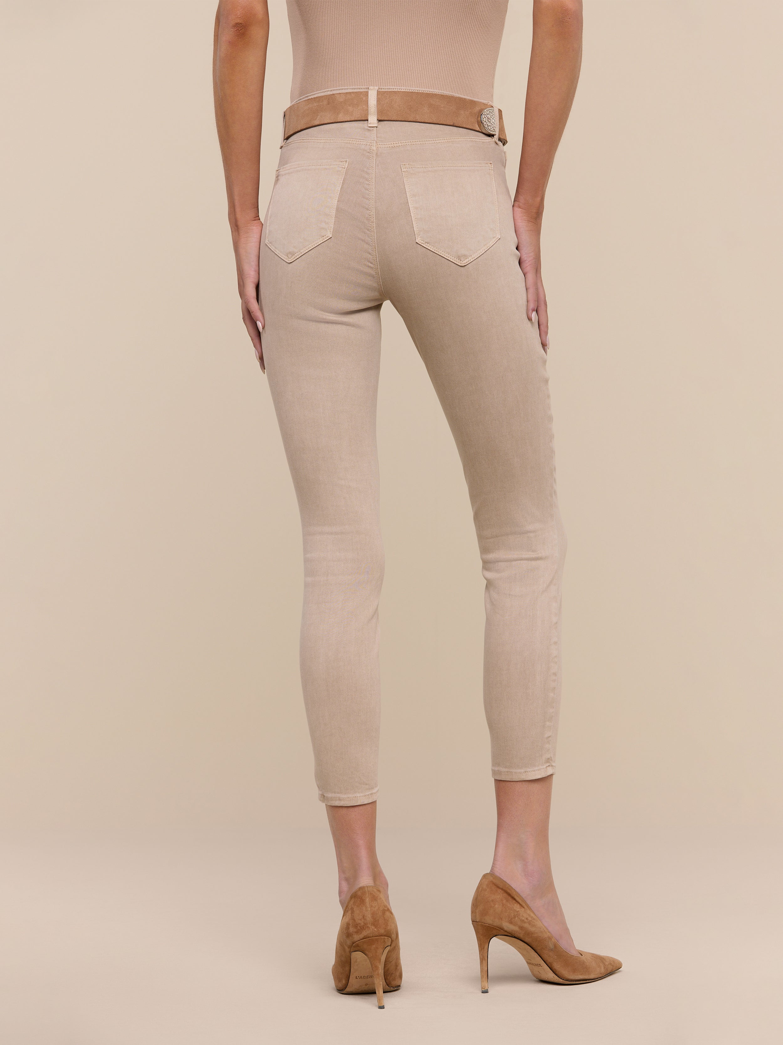 A person stands facing away against a plain white background, wearing Margot Cropped Skinny Jeans in beige stretch denim, styled with a matching belt and tan high-heeled shoes.