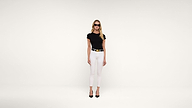 A woman wearing black sunglasses, a black fitted top, white pants, a black belt with a gold buckle, and black heels stands against a plain white background.