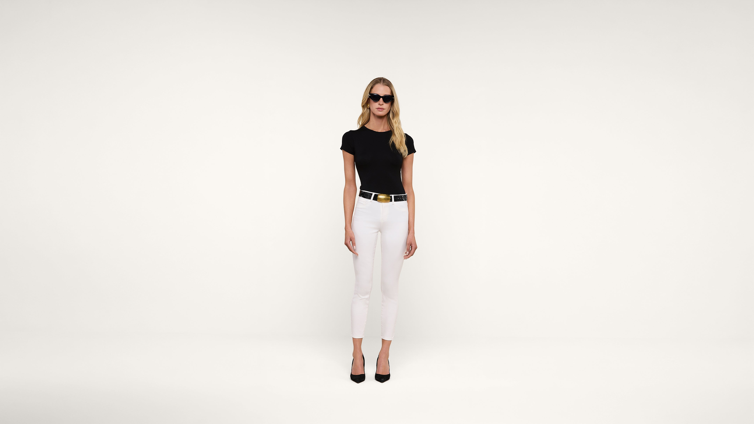 A woman wearing black sunglasses, a black fitted top, white pants, a black belt with a gold buckle, and black heels stands against a plain white background.
