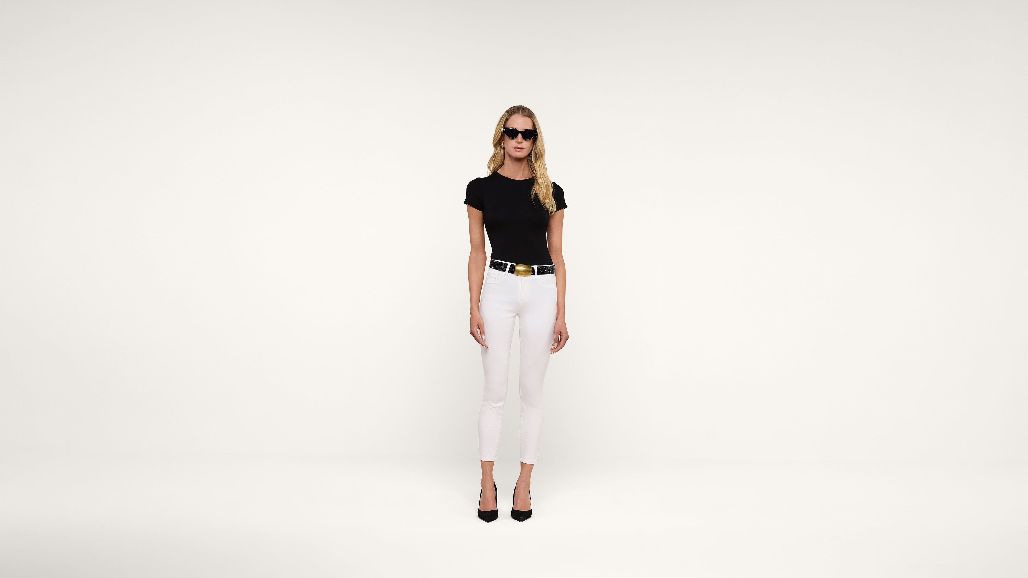 A woman wearing black sunglasses, a black fitted top, white pants, a black belt with a gold buckle, and black heels stands against a plain white background.