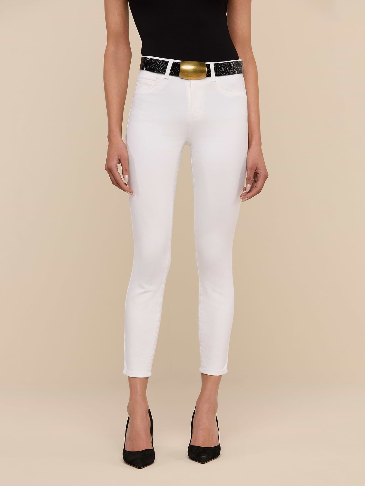 A woman stands against a beige background wearing a black sleeveless top, Margot Cropped Skinny Jean in white stretch denim, a black belt with a large gold buckle, and black pointed-toe heels. Her face is not visible.
