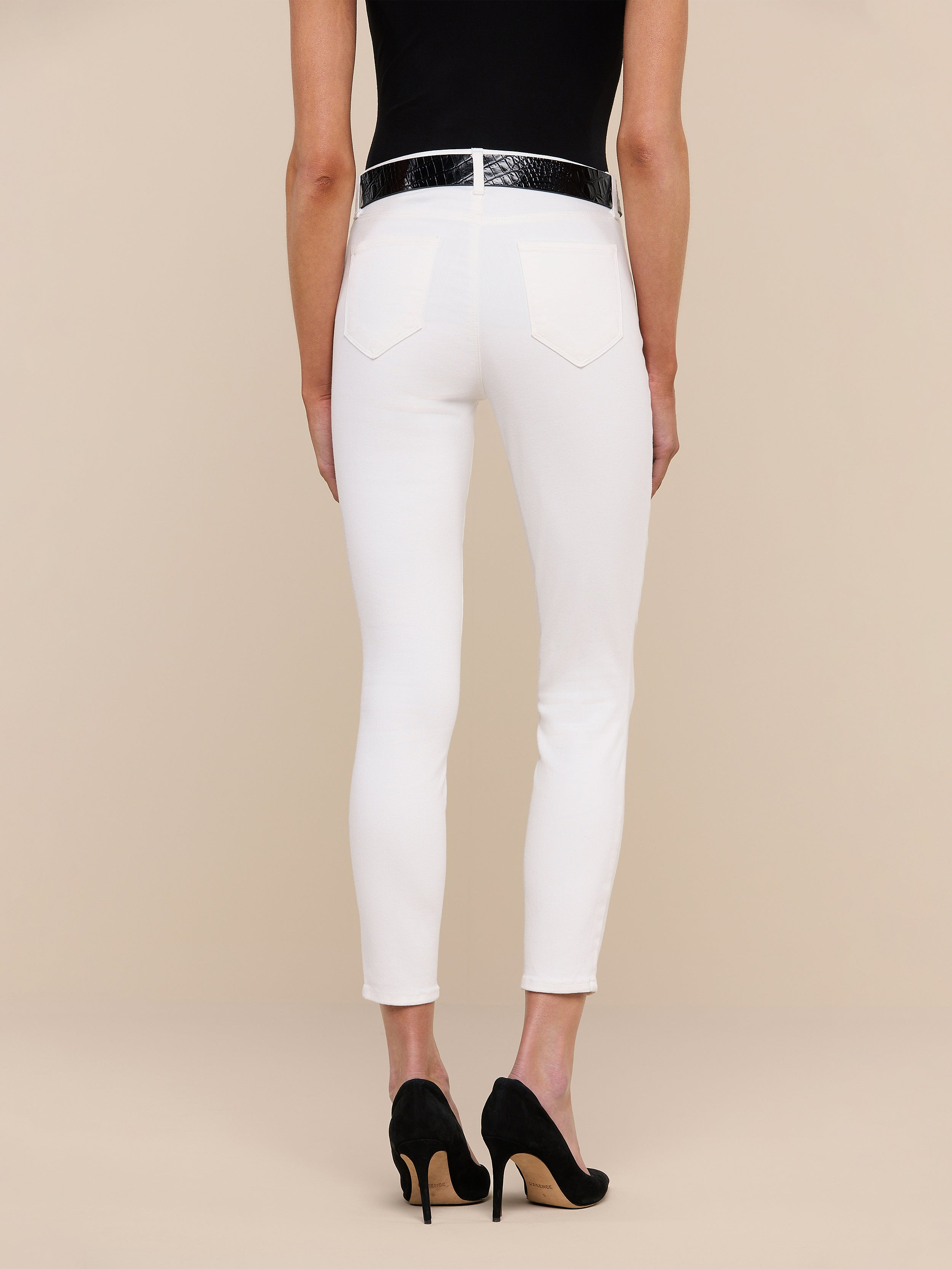 A person stands against a plain white background, shown from the back, wearing a black top and the Margot Cropped Skinny Jean in high-rise skinny white stretch denim with back pockets, paired with a black belt and black high-heeled shoes.