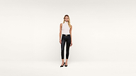 A woman stands against a plain white background, wearing a sleeveless white top, black leather pants, and black heels. She has long blond hair and is modeling with one hand at her side and the other slightly forward.