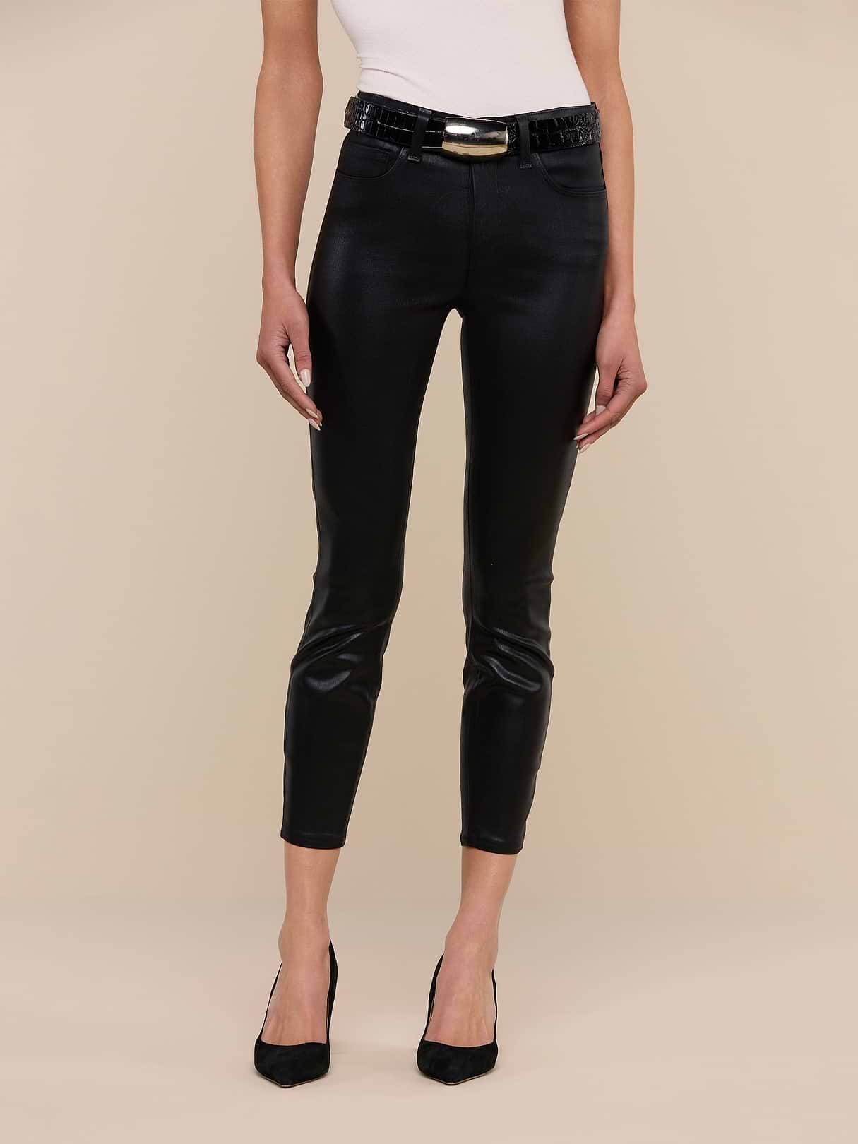A person wearing a white sleeveless top, Margot Coated Cropped Skinny Jeans with a contoured waistband and shiny belt, and black pointed-toe heels, standing against a neutral beige background.