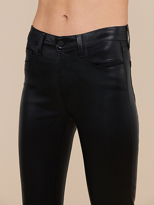 A close-up of a person wearing the Margot Coated Cropped Skinny Jean, a high-waisted black faux leather pant with tapered legs, front and side pockets, belt loops, and a button closure against a plain beige background.