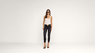 A woman wearing sunglasses, a white tank top, black skinny jeans, and heels stands confidently against a plain white background.