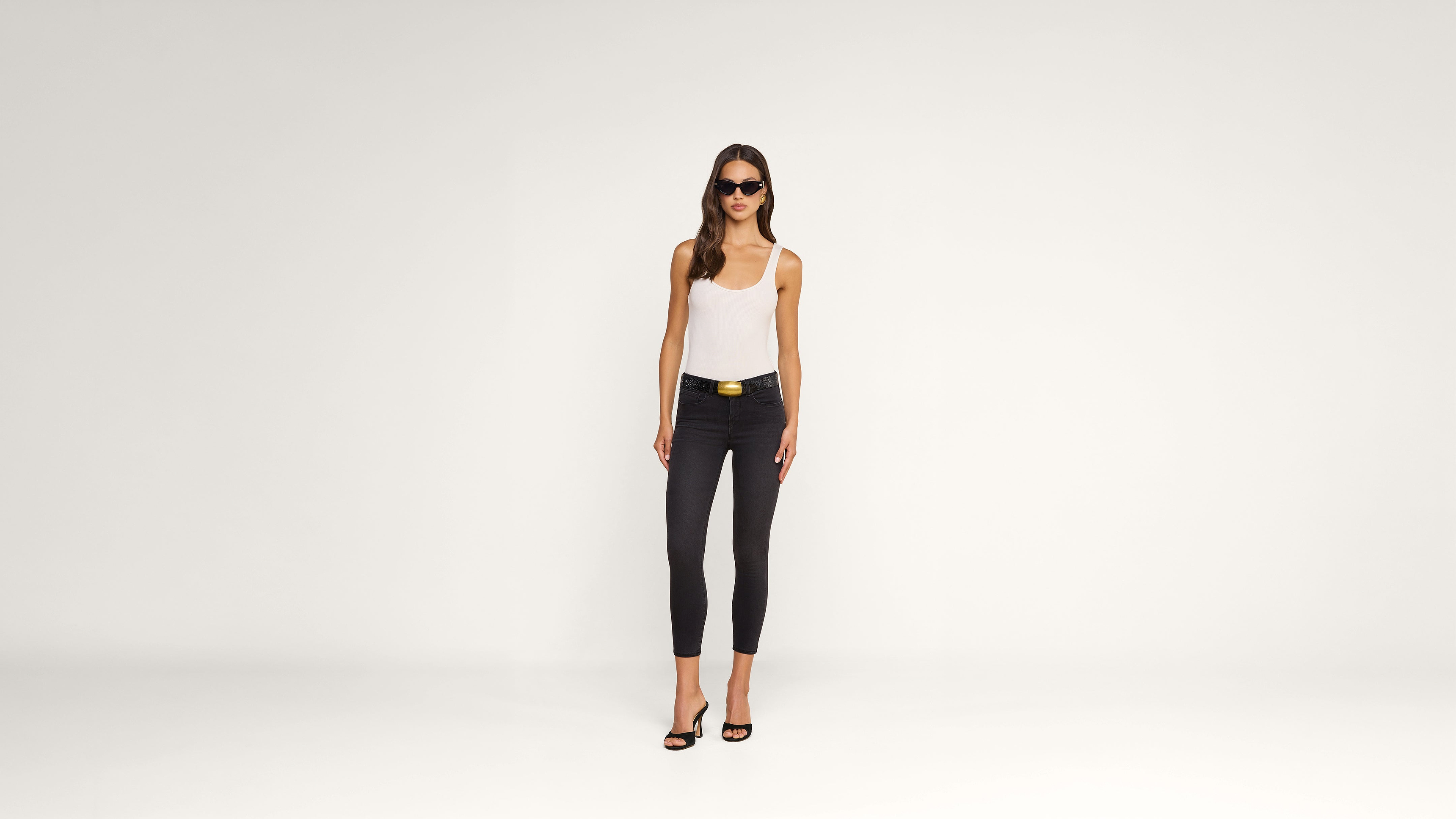 A woman wearing sunglasses, a white tank top, black skinny jeans, and heels stands confidently against a plain white background.