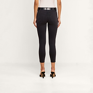 A woman stands facing away, wearing a white top and the Margot Cropped Skinny Jean—black high-rise skinny jeans with a contoured waistband—styled with a black belt and black heeled sandals against a plain light background.