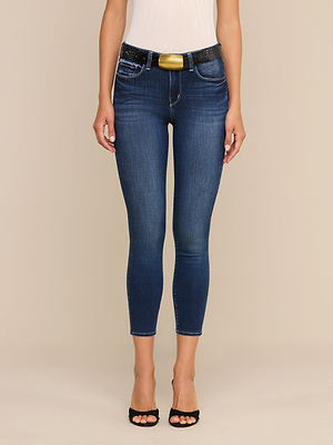 A woman wears Margot Cropped Skinny Jeans in dark blue premium stretch denim with recycled fiber, styled with a black glittery belt with a gold buckle, white sleeveless top, and black open-toe heels against a beige background.