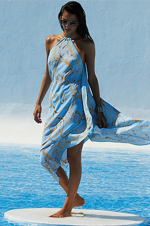 A woman in sunglasses stands barefoot near a pool, wearing a light blue, halter-neck, flowing dress with gold chain patterns. The background and floor are shades of blue, evoking a summery, tropical setting.