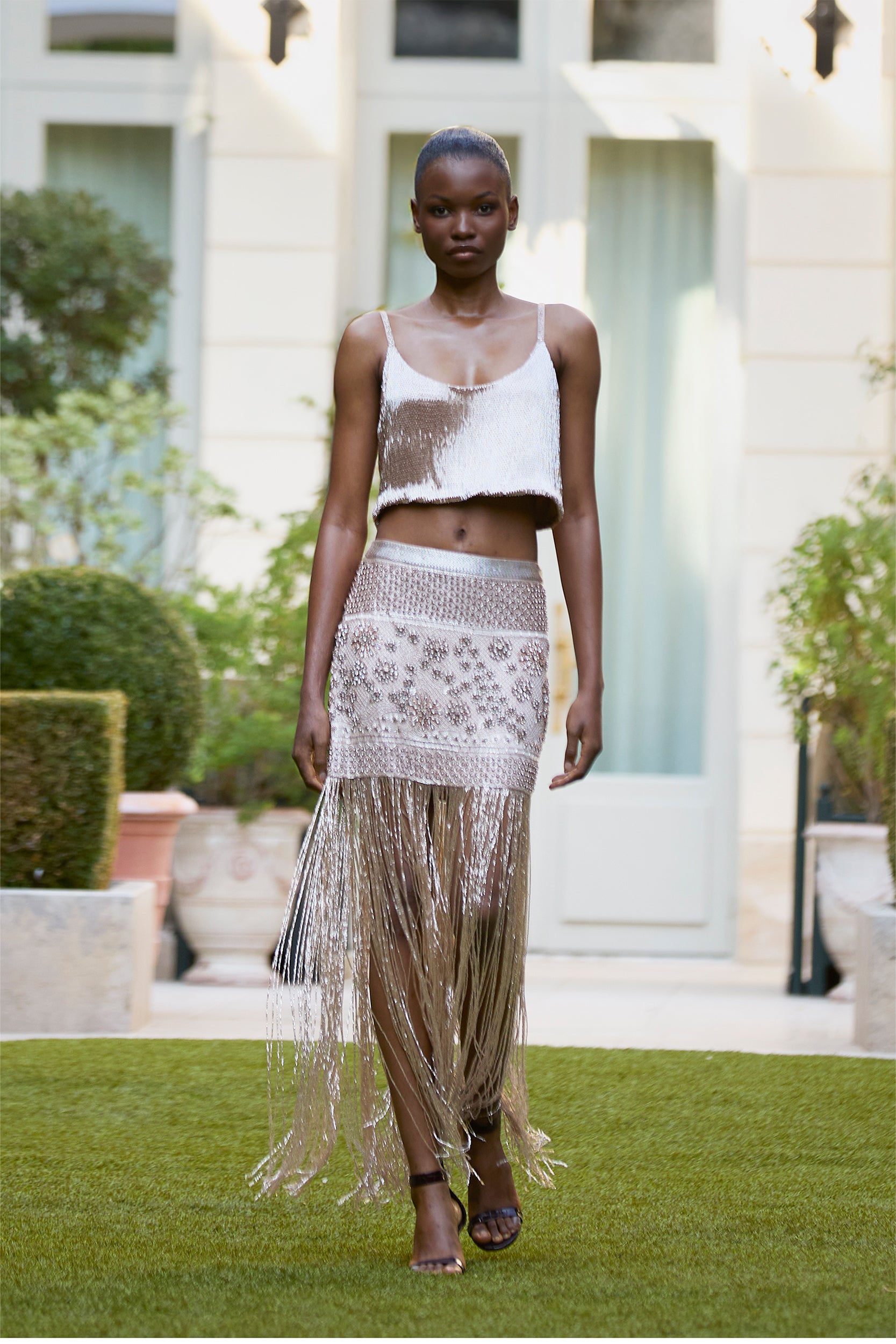 A model walks outside in a silver fringe crop top and matching sheer skirt with beaded details and long metallic fringe, set against lush greenery and a white building.