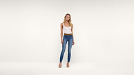 A woman stands in a white studio wearing a white tank top, blue jeans with a brown belt, and tan high heels. She looks at the camera with a neutral expression.