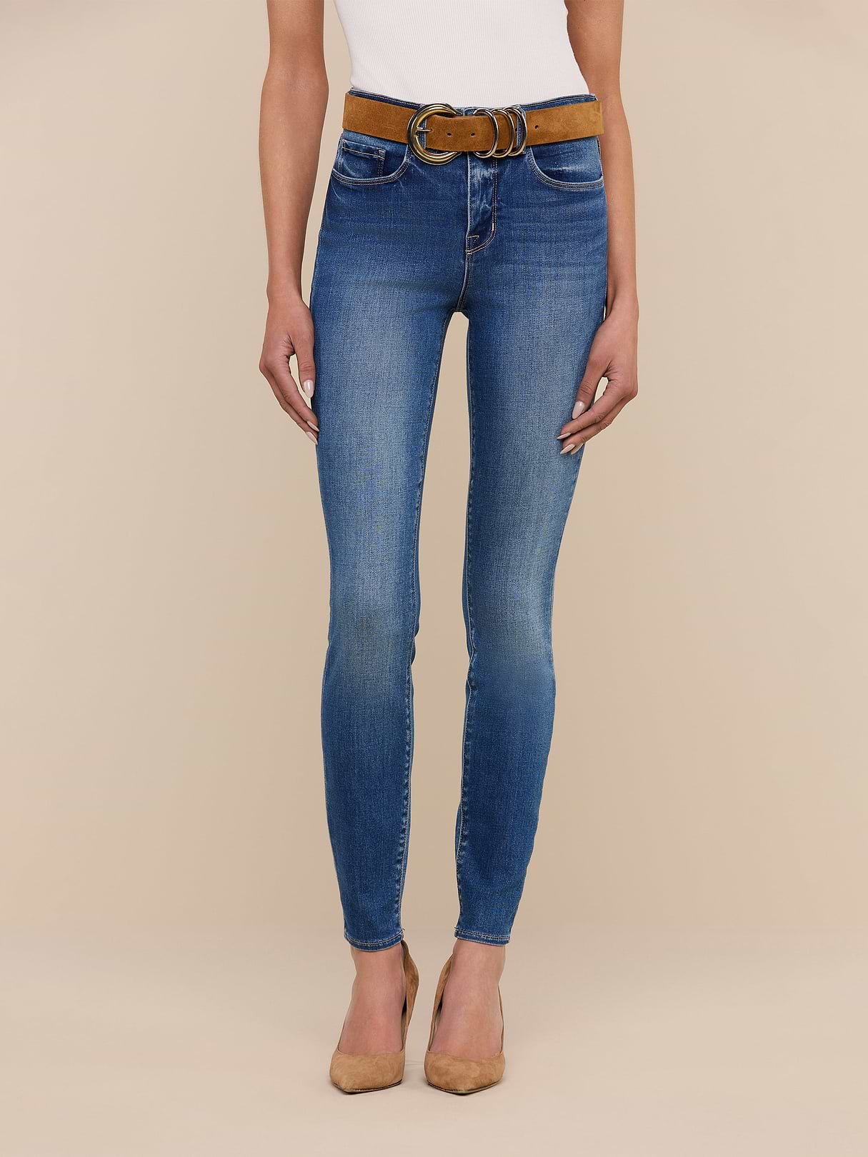Model showcases the Marguerite Skinny Jean, a high-rise blue skinny fit made from stretch denim, paired with a brown belt and tan pointed-toe heels. Only the body from shoulders down is visible against a beige background.