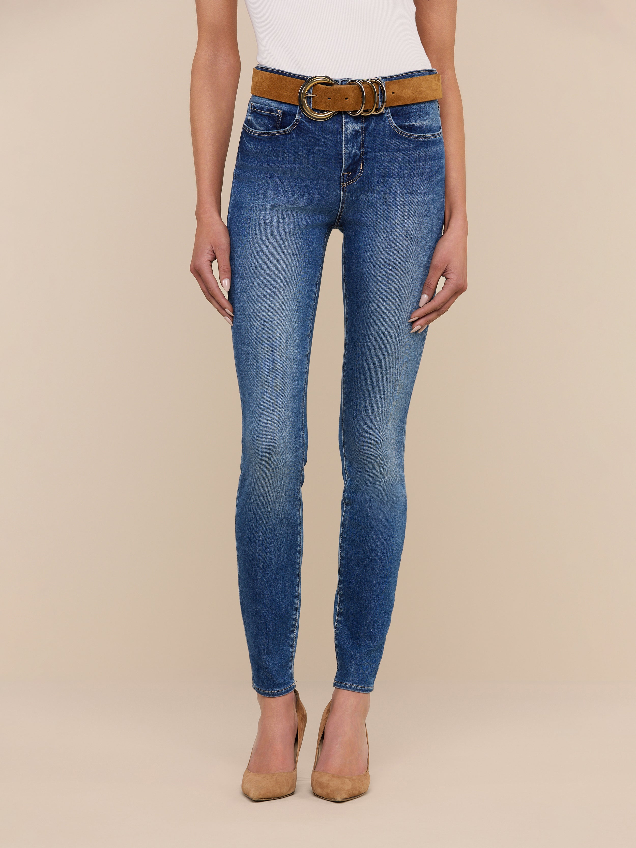 Model showcases the Marguerite Skinny Jean, a high-rise blue skinny fit made from stretch denim, paired with a brown belt and tan pointed-toe heels. Only the body from shoulders down is visible against a beige background.