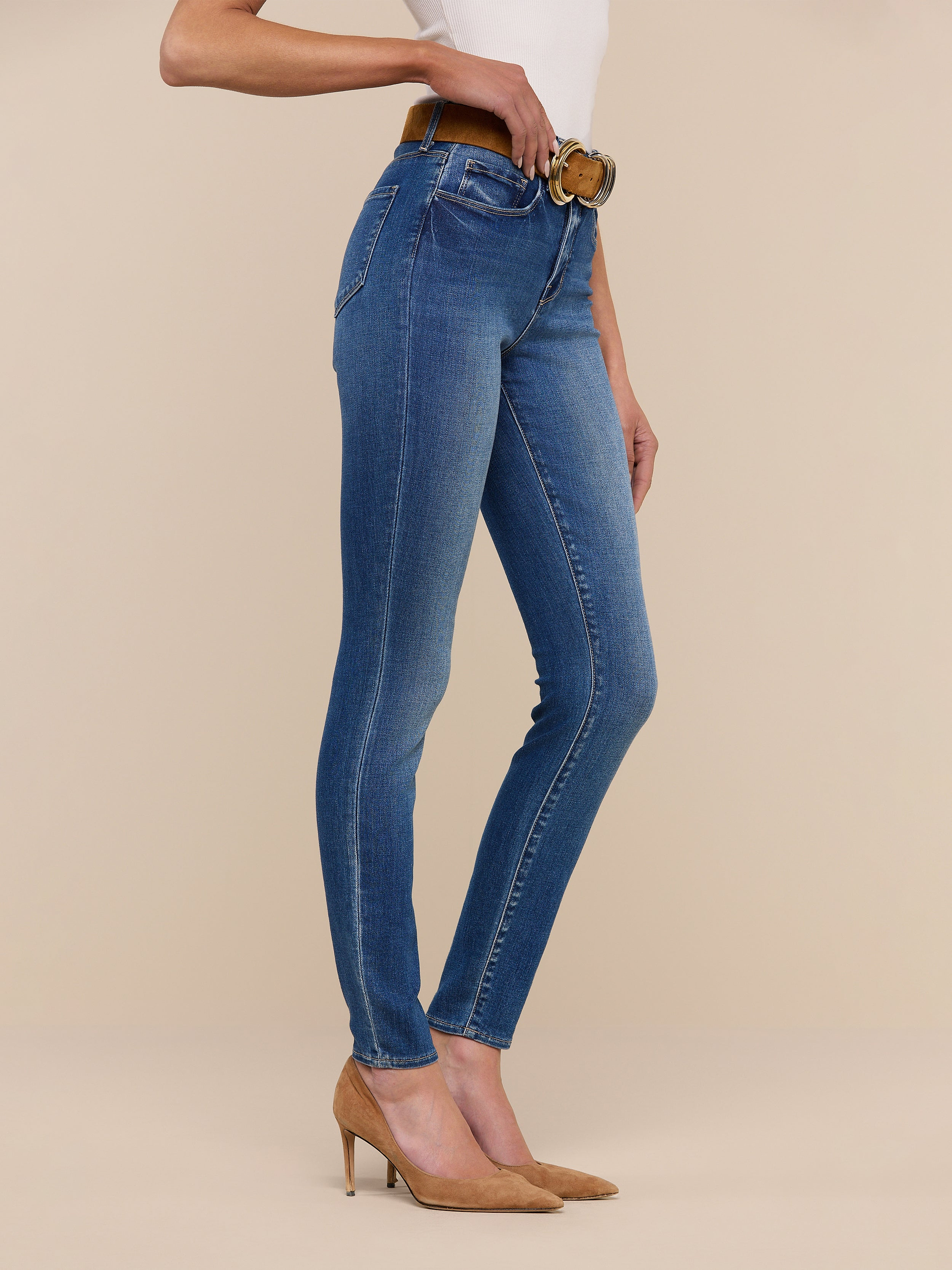 A person stands sideways against a beige background, showing only their lower half while wearing the Marguerite Skinny Jean with a brown belt, white top, and tan heels.