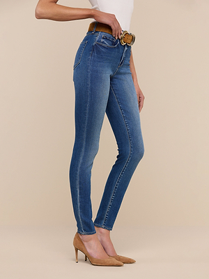 A person stands sideways against a beige background, showing only their lower half while wearing the Marguerite Skinny Jean with a brown belt, white top, and tan heels.