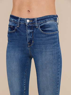 A close-up shows a person wearing the Marguerite Skinny Jean in high-rise blue stretch denim, highlighting the waist, pockets, and upper thighs. This skinny style features a button closure and visible stitching against a neutral background.