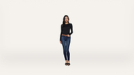 A woman with long dark hair stands against a white background, wearing a black long-sleeve shirt, blue jeans, a brown belt, and black shoes. She is facing forward with one hand on her hip.