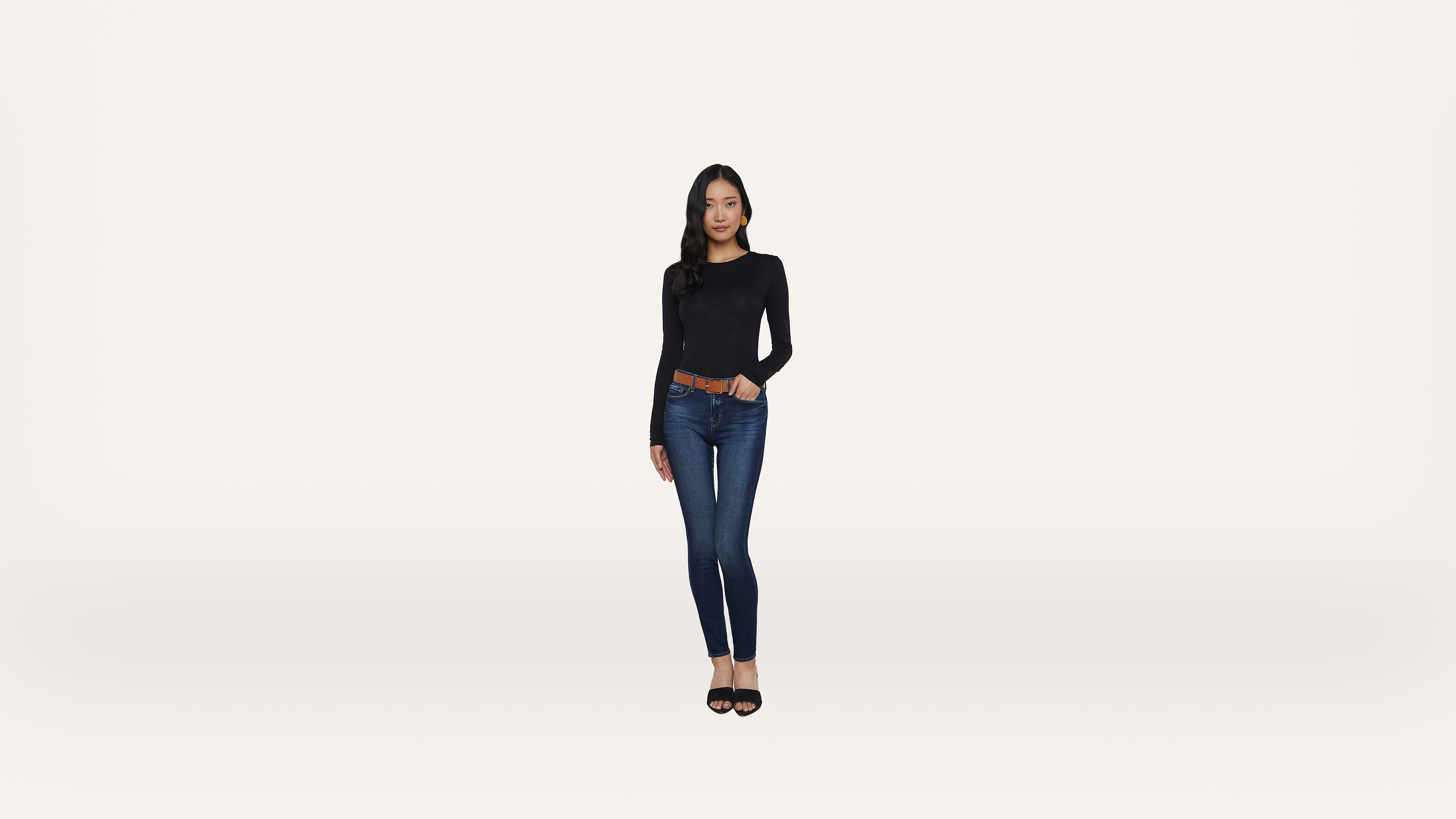 A woman with long dark hair stands against a white background, wearing a black long-sleeve shirt, blue jeans, a brown belt, and black shoes. She is facing forward with one hand on her hip.