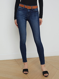A person wearing the Marguerite Skinny Jean in dark blue—crafted from premium stretch denim—paired with a brown belt, a black long-sleeve top, and black open-toe shoes stands against a plain white background.
