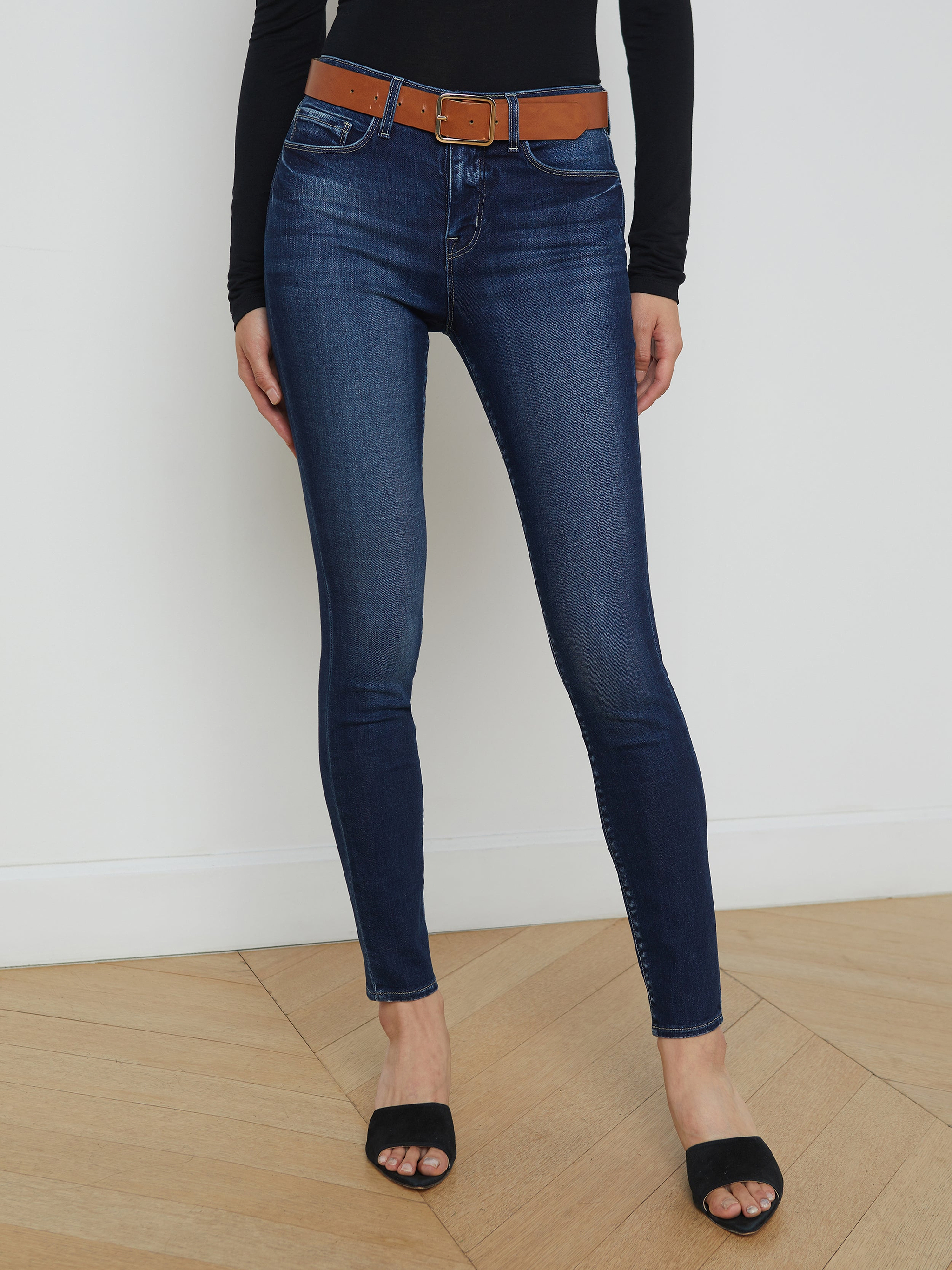 A person wearing the Marguerite Skinny Jean in dark blue—crafted from premium stretch denim—paired with a brown belt, a black long-sleeve top, and black open-toe shoes stands against a plain white background.