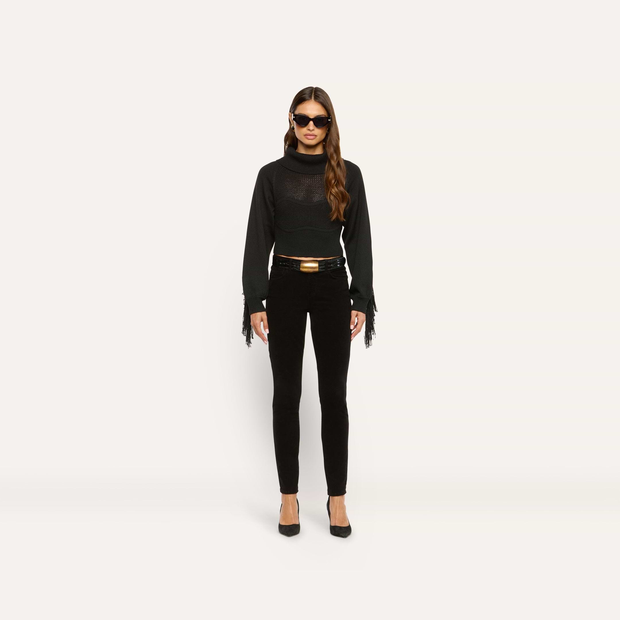 A model wears high-rise black Marguerite Corduroy Skinny Pant with a black fringe-sleeved sweater, a black belt with a gold buckle, and black pumps.