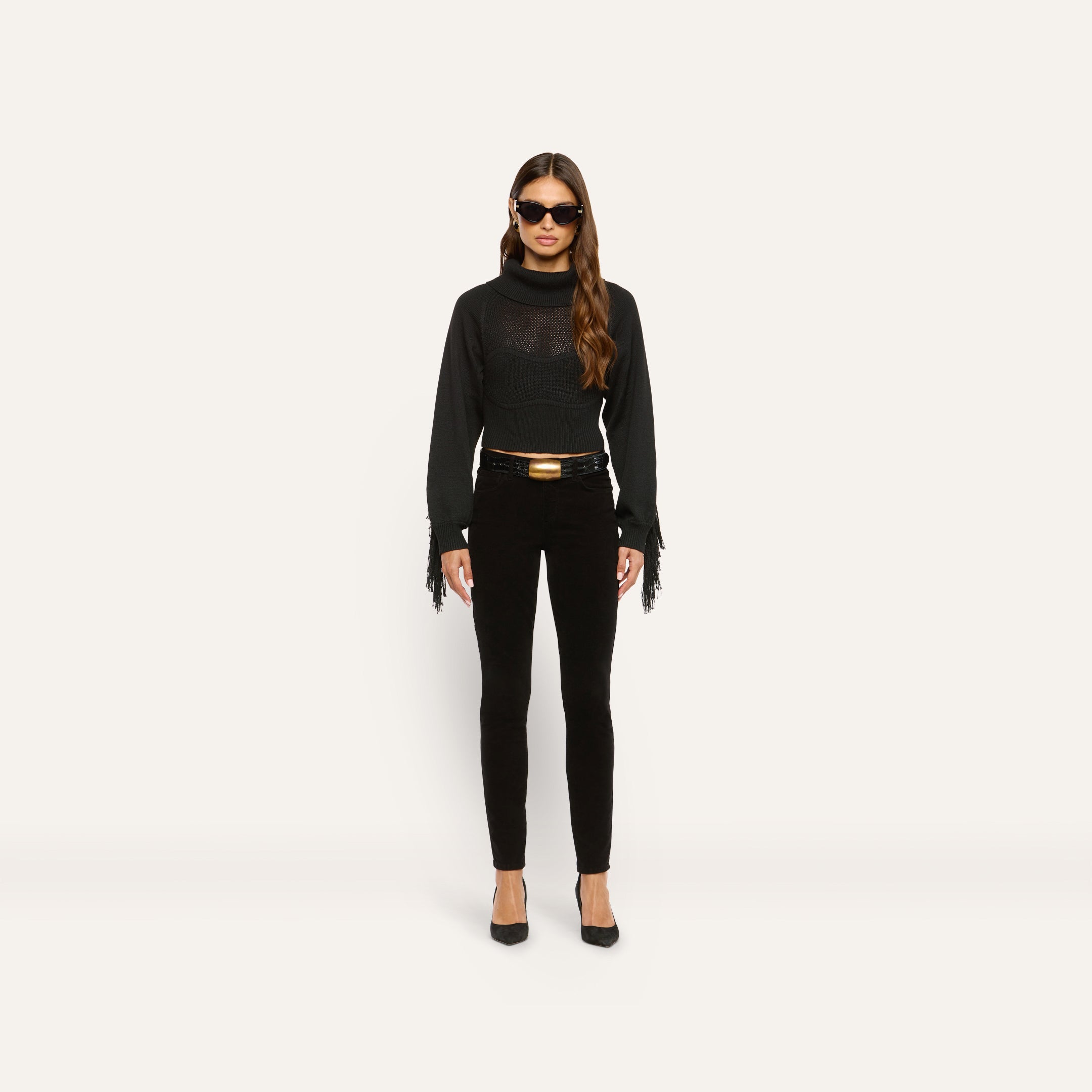 A model wears high-rise black Marguerite Corduroy Skinny Pant with a black fringe-sleeved sweater, a black belt with a gold buckle, and black pumps.