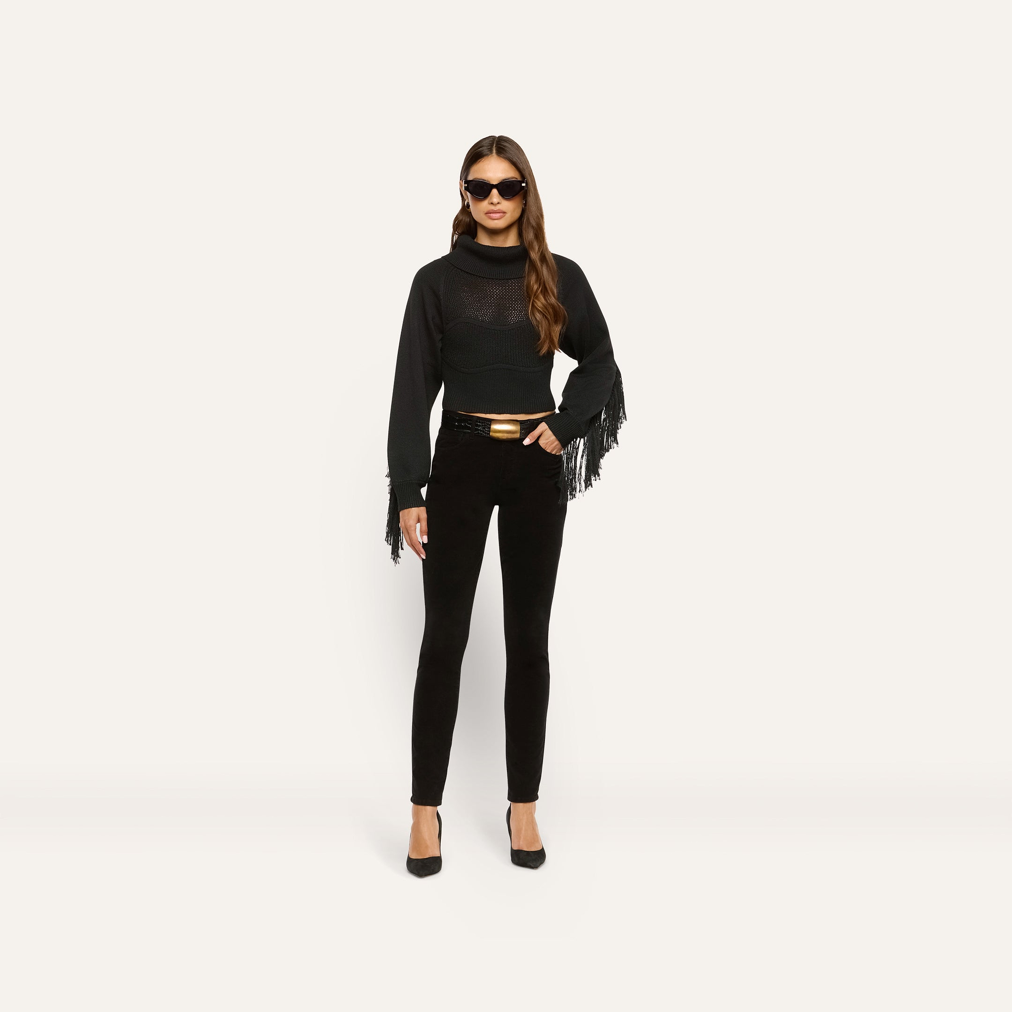 A woman wearing a black long-sleeve fringe sweater, a belt with a gold buckle, and Marguerite Corduroy Skinny Pant in black.