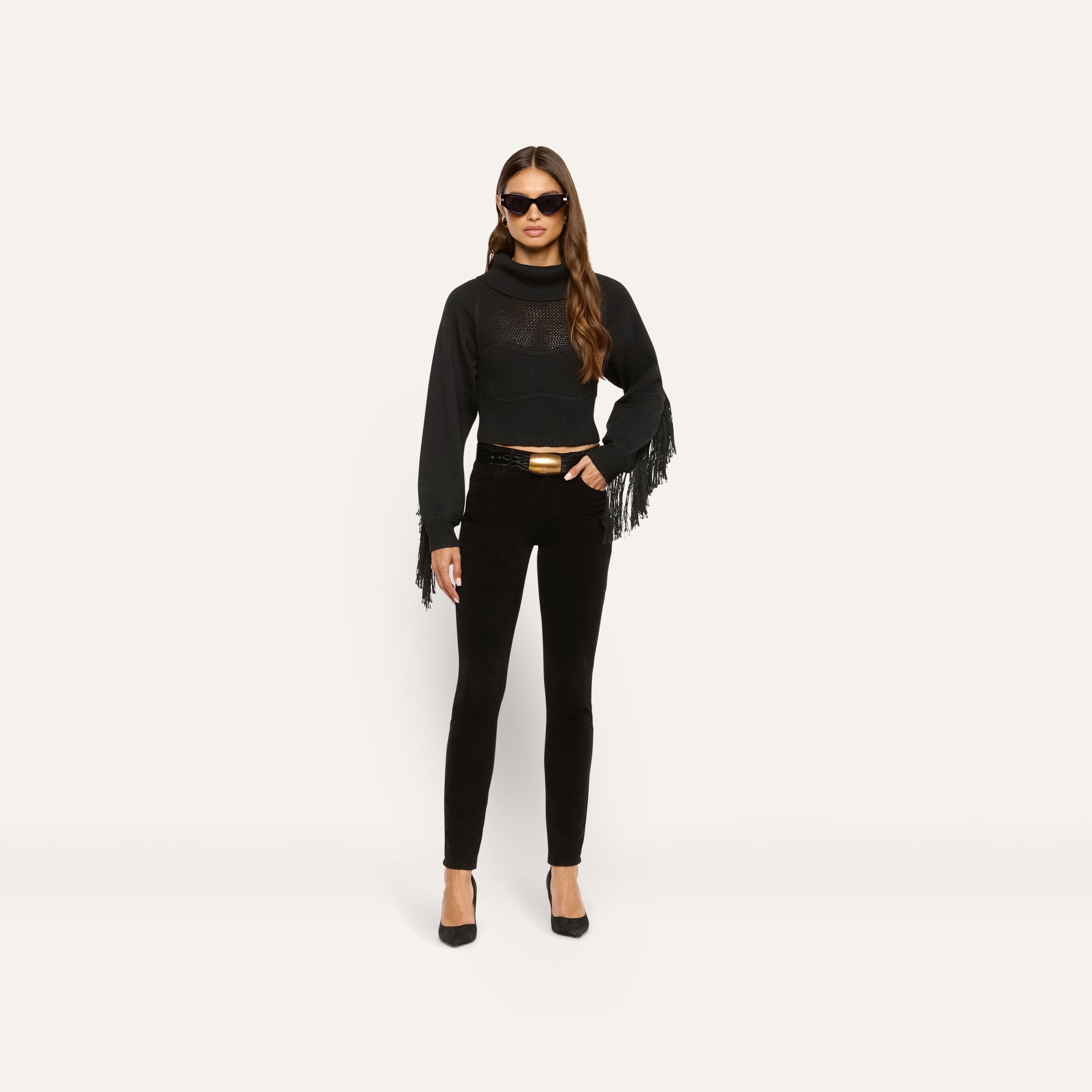 A woman wearing a black long-sleeve fringe sweater, a belt with a gold buckle, and Marguerite Corduroy Skinny Pant in black.