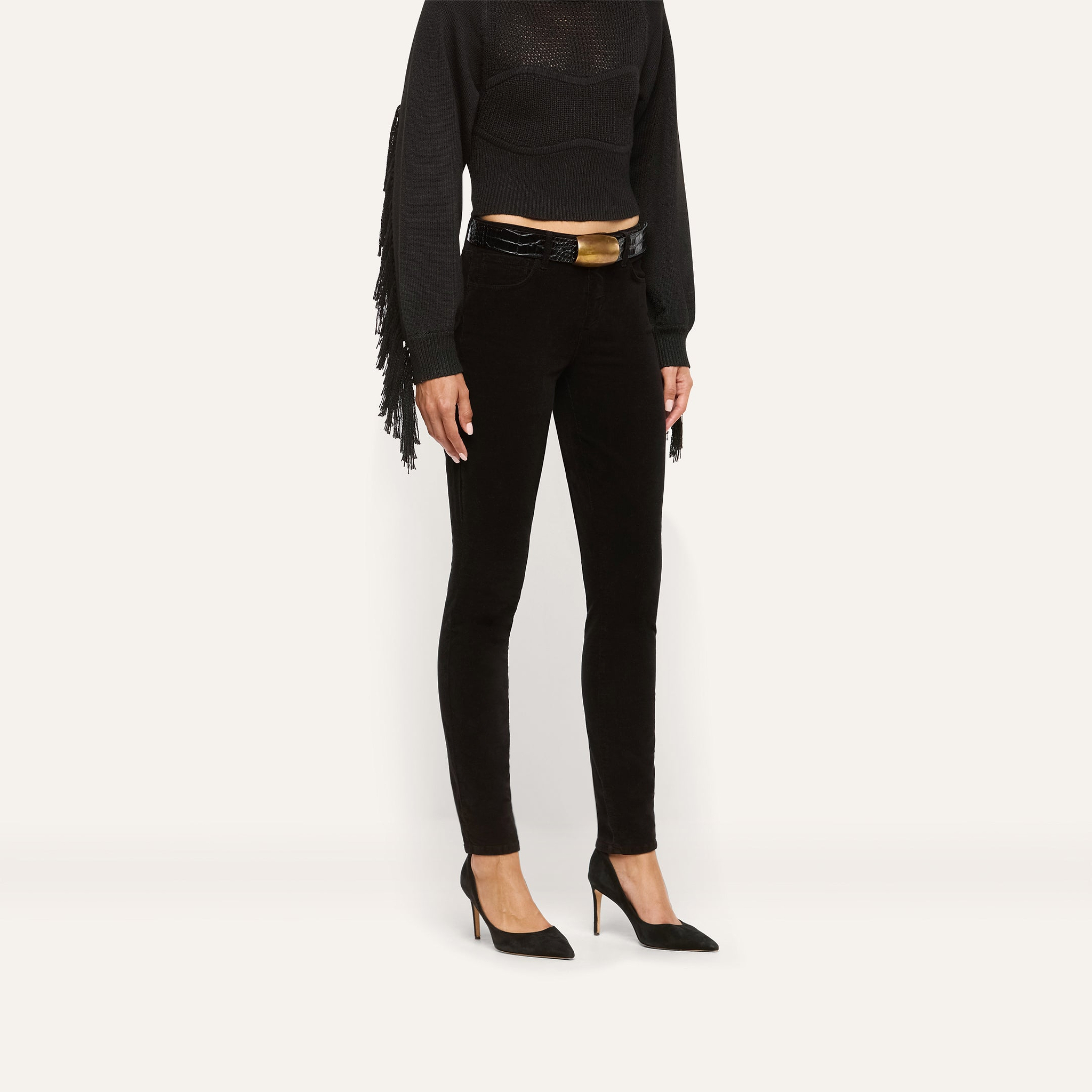 Marguerite Corduroy Skinny Pant in high-rise black corduroy with an embossed belt featuring a large gold buckle, worn with a black fringed top and black pointed-toe heels.
