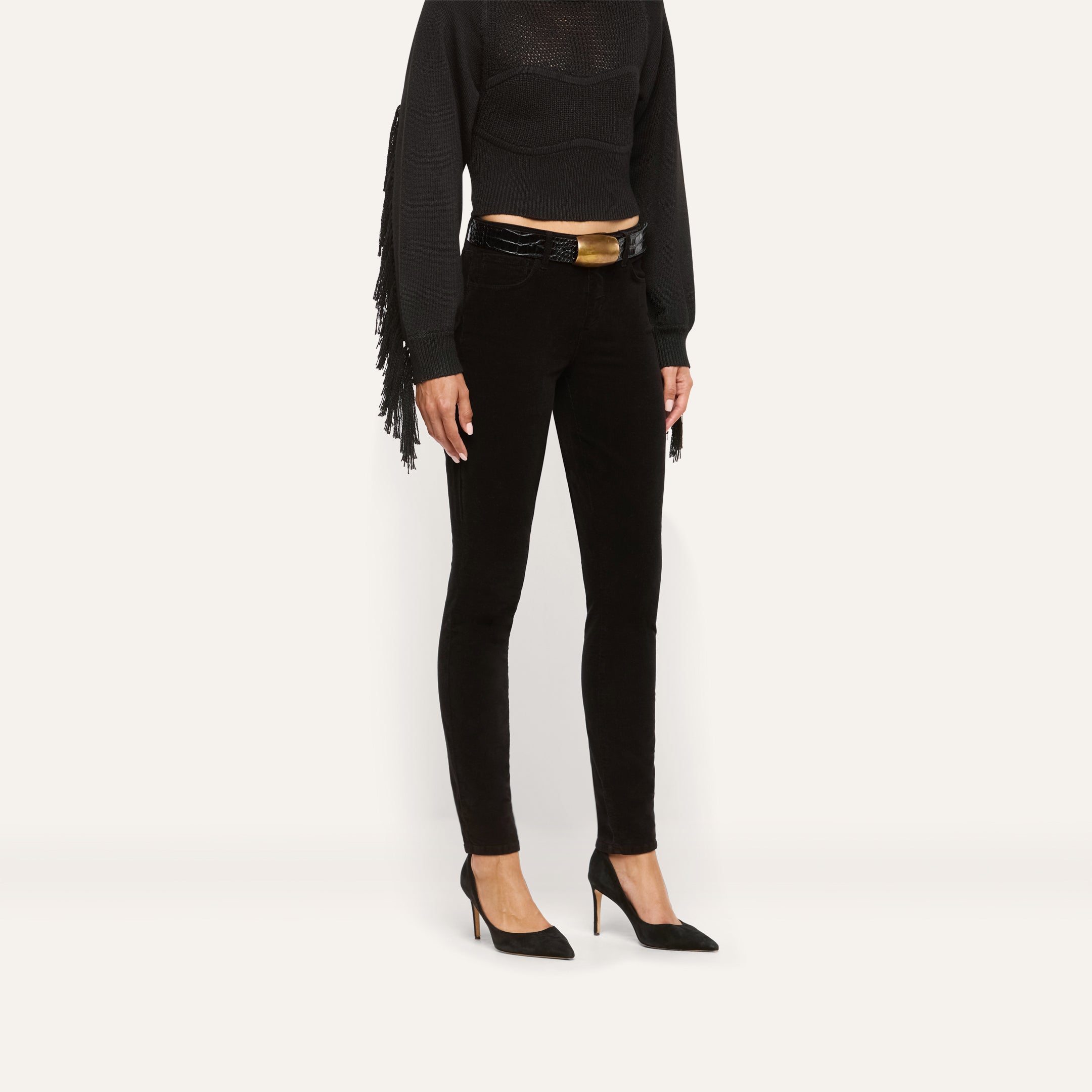 Marguerite Corduroy Skinny Pant in high-rise black corduroy with an embossed belt featuring a large gold buckle, worn with a black fringed top and black pointed-toe heels.