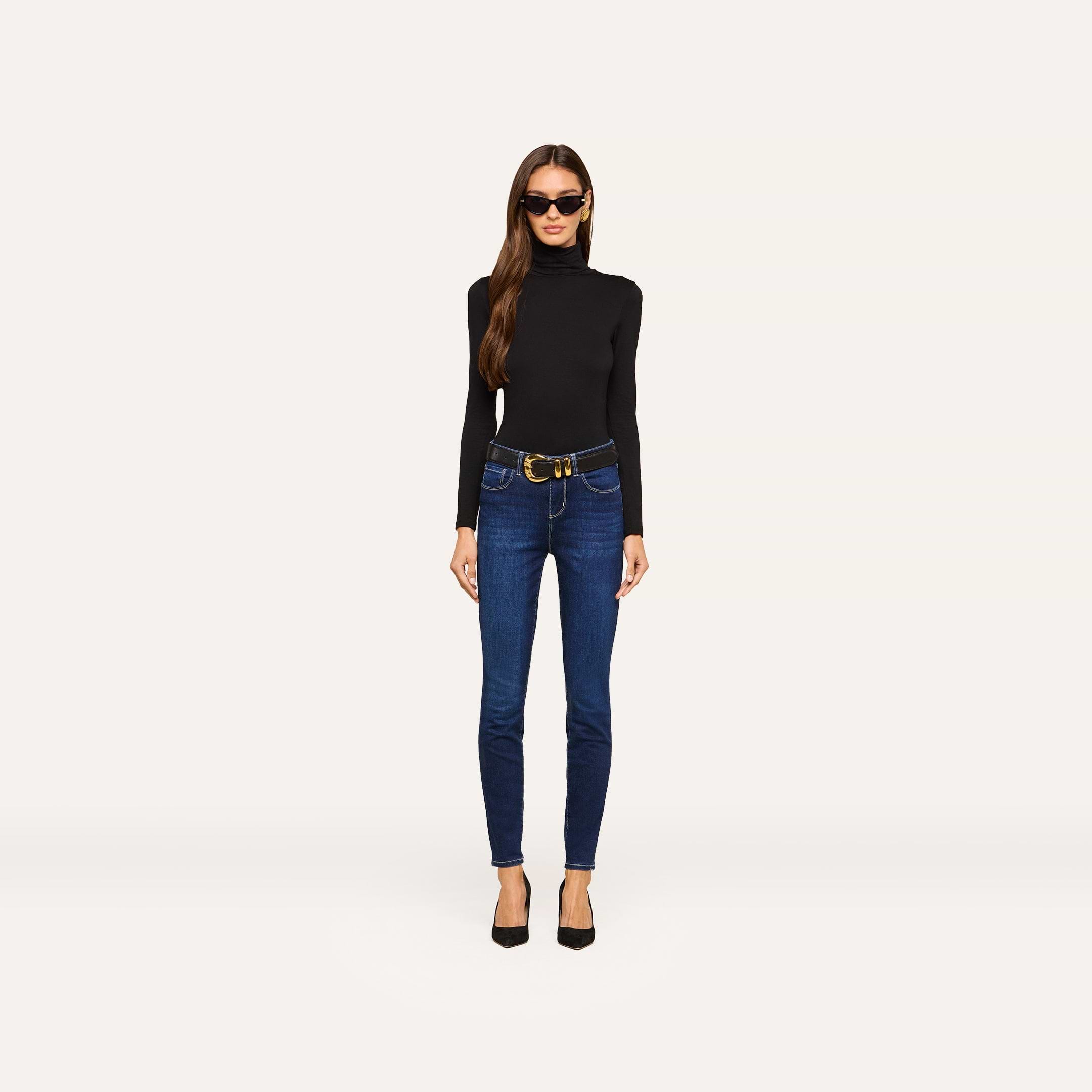 Wearing sunglasses, a black turtleneck sweater, black heels, and the Marguerite Skinny Jean with a belt, a woman stands against a plain, light background.