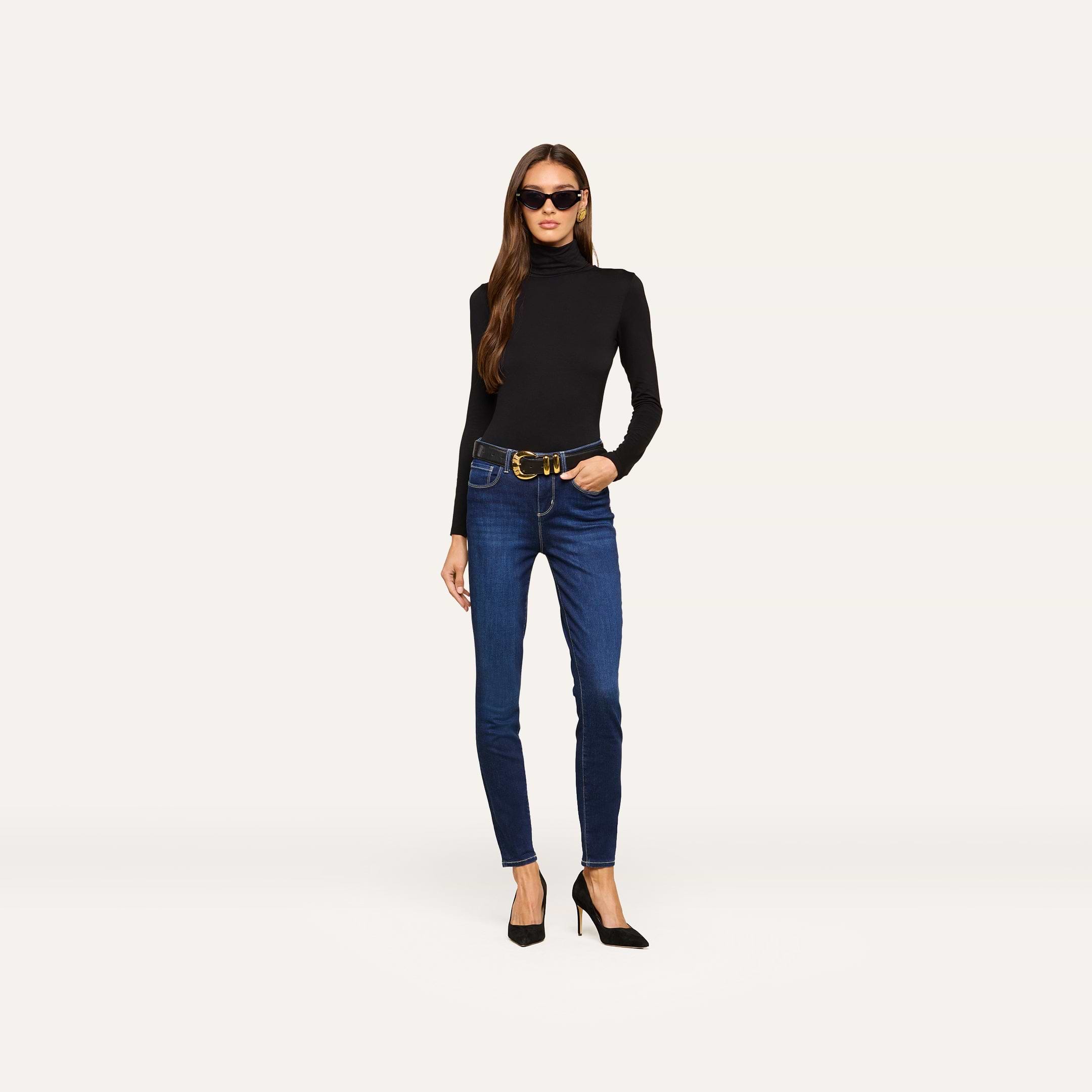 A woman wearing sunglasses, a black long-sleeve turtleneck, a belt, and black high heels stands against a plain light background. She completes her outfit with the Marguerite Skinny Jean—blue high rise skinny jeans crafted from stretch denim.