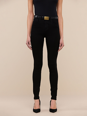 A person wearing a black sleeveless top, Marguerite Skinny Jean in black with a matching belt featuring a gold buckle, and black pointed-toe heels stands against a plain beige background.