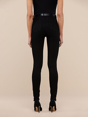 A person faces away from the camera, wearing Marguerite Skinny Jean—high-waisted black skinny jeans made from high-recovery fabric—paired with a black belt, top, and high-heeled shoes against a beige background.