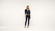 A woman stands facing forward against a plain white background, wearing a dark blue blouse, dark skinny jeans, tan heels, and a tan belt, with one hand resting by her side and the other slightly bent.