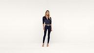 A woman stands against a plain white background, wearing a dark blue blouse, matching fitted pants, a brown belt, and beige heels. She has long blonde hair and a neutral expression, creating an effortlessly chic look.